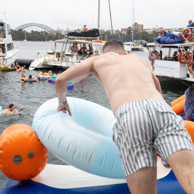 A man in striped shorts climbs onto a large inflatable float near a dock. Boats and other inflatables are in the water, with a cityscape and bridge visible in the background. The scene is lively, suggesting a festive gathering.