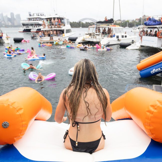 A person in a black swimsuit sits on an inflatable slide, facing a lively water scene with boats and people on floaties. The background features a city skyline and overcast sky, suggesting a festive gathering on the water.