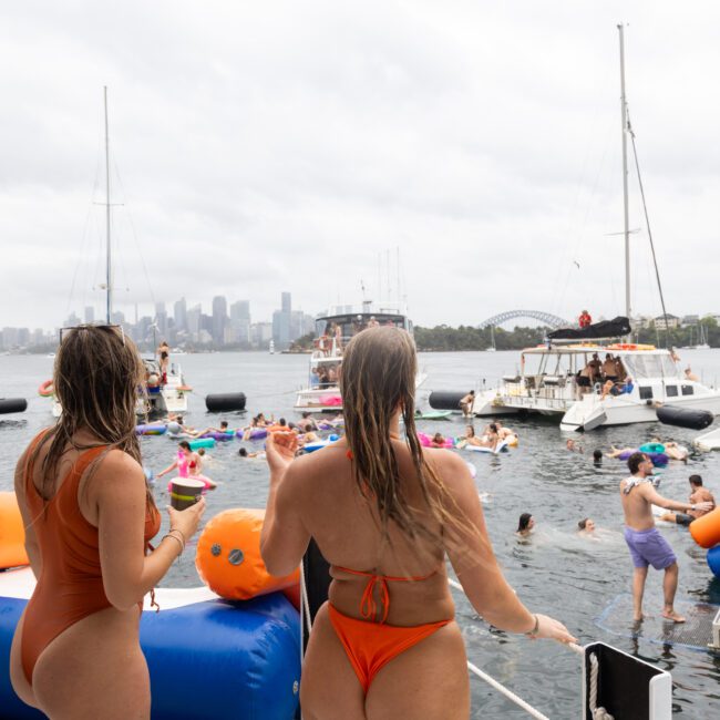 Two women in orange swimsuits stand on a boat overlooking a lively harbor party with people on floats and boats. The city skyline is visible in the distance under a cloudy sky.