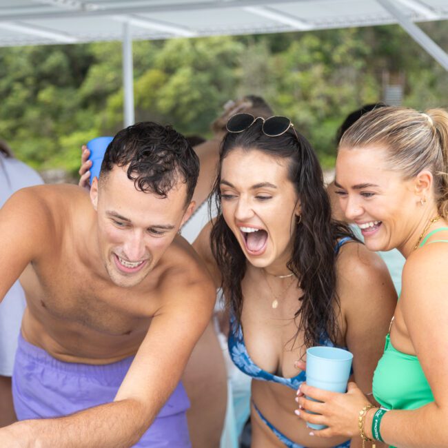 A group of people in swimsuits enjoy themselves on a boat. Three people in the foreground smile and laugh, one holding a light blue cup. Lush greenery is in the background.