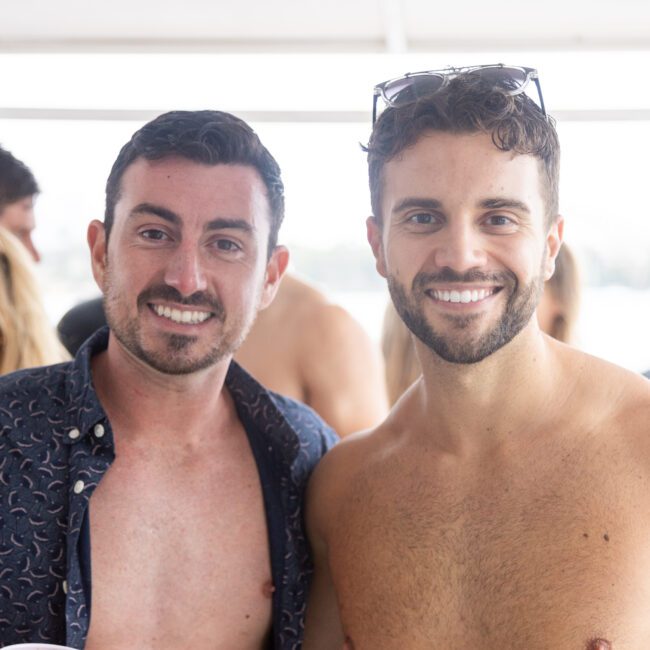 Two men smiling at the camera on a boat. One is wearing an open dark shirt, and the other is shirtless. They appear to be enjoying a sunny day on the water with other people in the background.