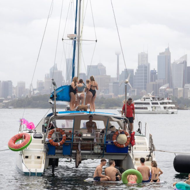 A group of people enjoying a day on a catamaran, with some standing on the deck and others swimming in the water. The city skyline is visible in the background under a cloudy sky. Inflatable rings float nearby.