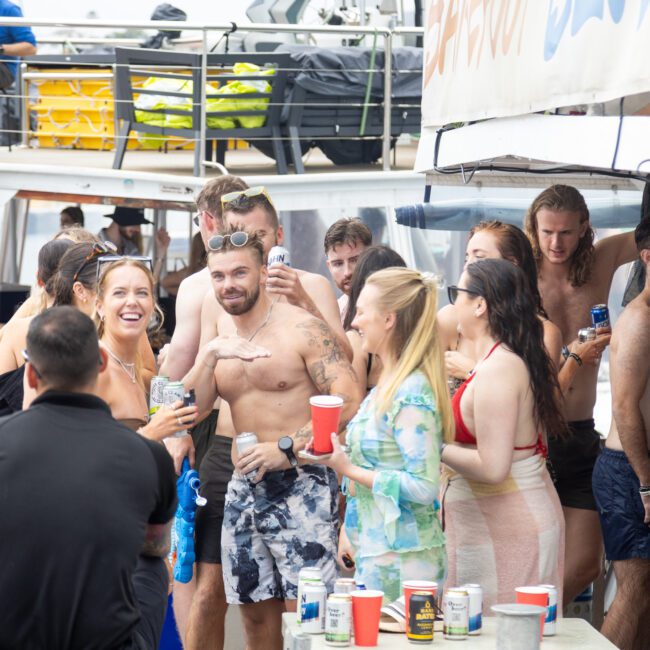 A lively group of people enjoying a party on a boat. They're wearing swimwear, holding drinks, and smiling. The setting is casual and festive, with beverages on a table and some individuals sitting and chatting.