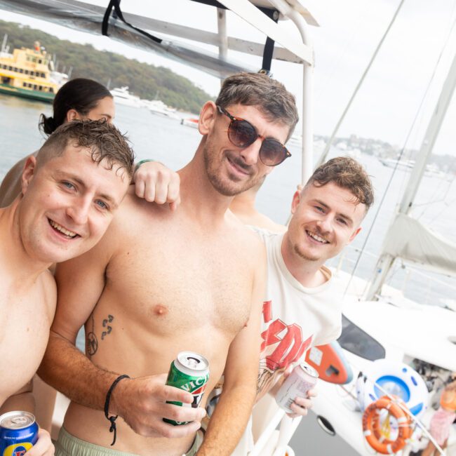 Three men with drinks pose shirtless on a boat, smiling at the camera. Behind them, a bay with other boats and a ferry is visible. The atmosphere is festive and relaxed, with a partly cloudy sky overhead.