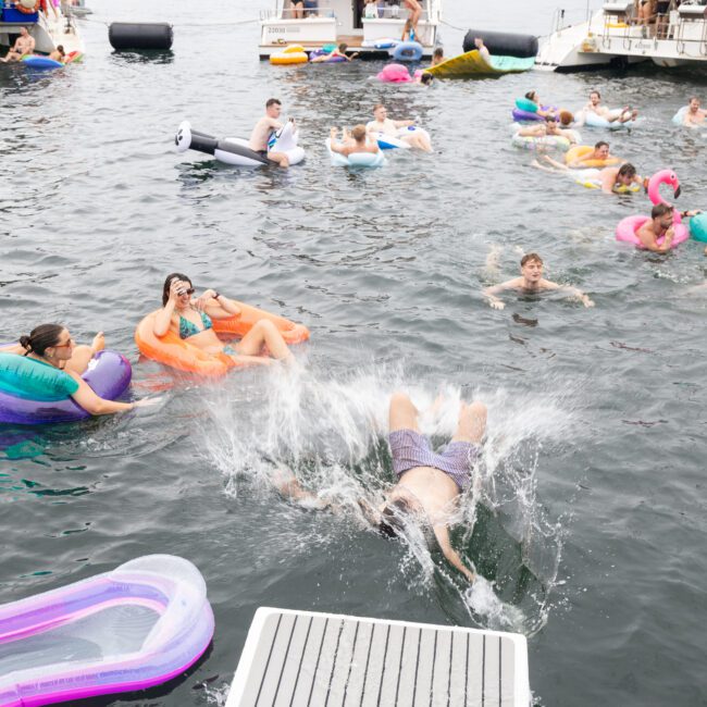 People enjoying a day on the water with colorful inflatable floats. In the foreground, a person is captured in mid-splash after jumping in. Boats and more people relaxing are visible in the background.