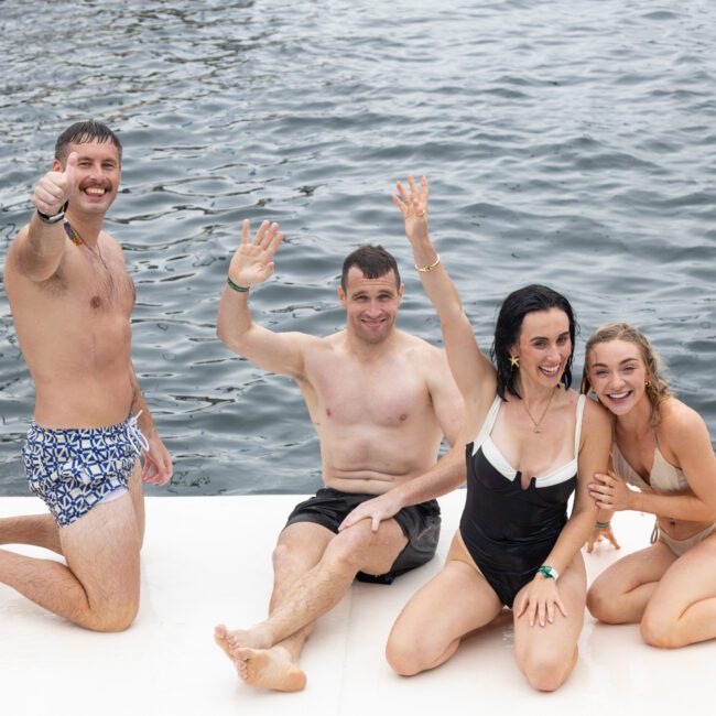 Four people in swimwear sit and kneel on a floating platform by the water. Three are waving, and one gives a thumbs-up. They appear to be having a fun time together. The background shows a calm body of water.