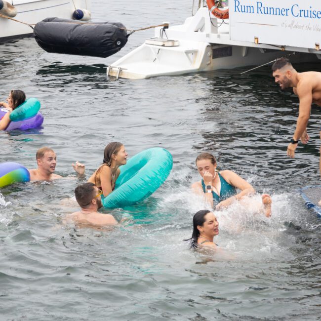 People are enjoying a swim near a boat. Some are floating on colorful inflatables while others are in the water. A shirtless man stands on a floating mat, about to jump. The sign on the boat reads "Rum Runner Cruises".