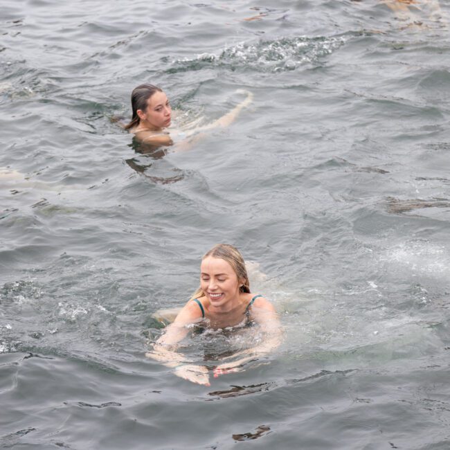 People swimming in a body of water, with a smiling woman in the foreground. The water ripples as they swim, and there are a couple more swimmers visible in the background. A purple floatation device is partially visible.