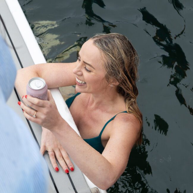 A person with blonde hair smiles while in the water, holding a can. They rest one arm on the edge of a boat. The water is dark and the person's nails are painted red. A logo in the corner reads "White Sand Beach.