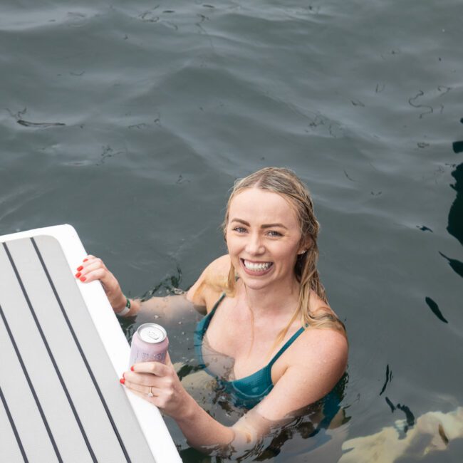 A woman in a green swimsuit is smiling while holding a can, swimming near the edge of a boat platform on a sunny day. The water is a deep shade of blue.