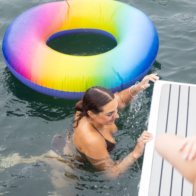 A woman in a black swimsuit is in the water, holding onto the edge of a dock. A colorful rainbow inflatable ring floats nearby. The water is calm, and another person's legs are visible in the foreground.