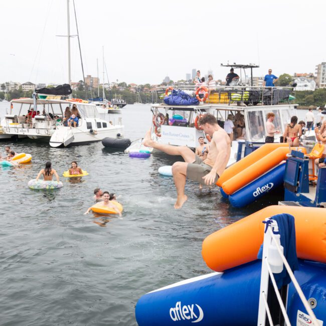 People enjoying a party on a boat with inflatable floats and slides. Some are swimming in the water, while others relax on the deck. Several other boats are anchored nearby, and a cityscape is visible in the background.