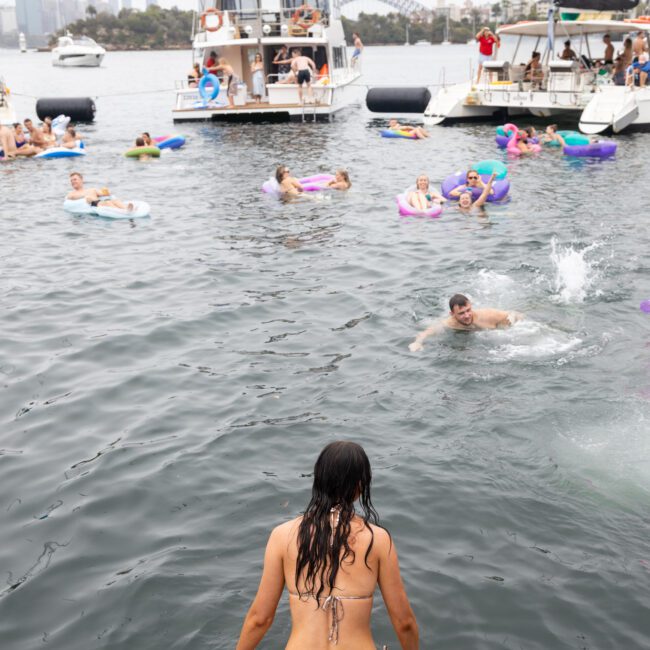 A woman in a bikini sits on a boat deck looking at people swimming and floating on inflatables in the water. Several boats and yachts are anchored nearby, and a city skyline is visible in the background.