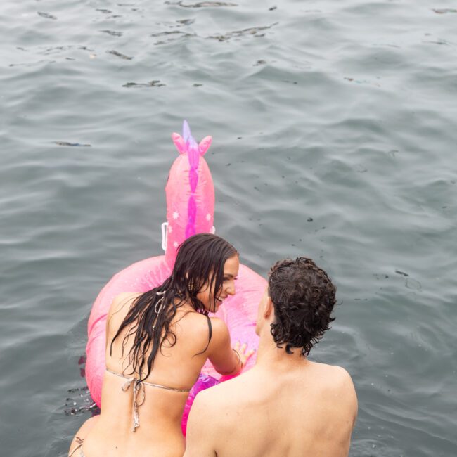 A woman and man sit on a dock by the water. The woman reaches for a large pink inflatable flamingo in the water. Both are in swimsuits, enjoying a relaxed moment at the lake.