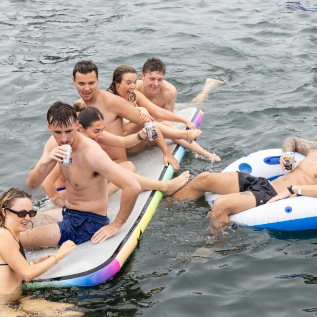 A group of people are enjoying themselves in the water. Some are sitting on a large inflatable board, while another person relaxes in a circular float. They are smiling and holding cans, suggesting a fun, social outing.