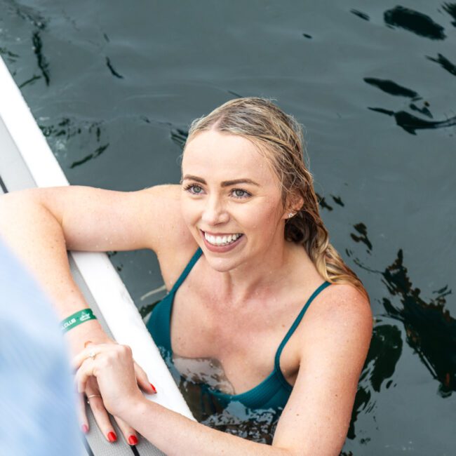 Woman in a green swimsuit smiling while holding the edge of a boat, partially submerged in water.