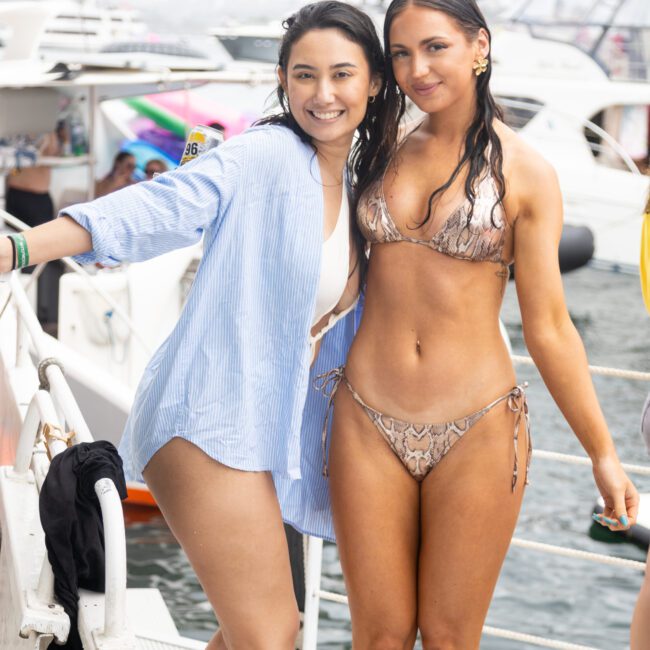 Two women smiling on a boat. One wears a blue shirt over a swimsuit, and the other is in a snakeskin-patterned bikini. Background includes other boats and people, suggesting a lively, sunny outing on the water.