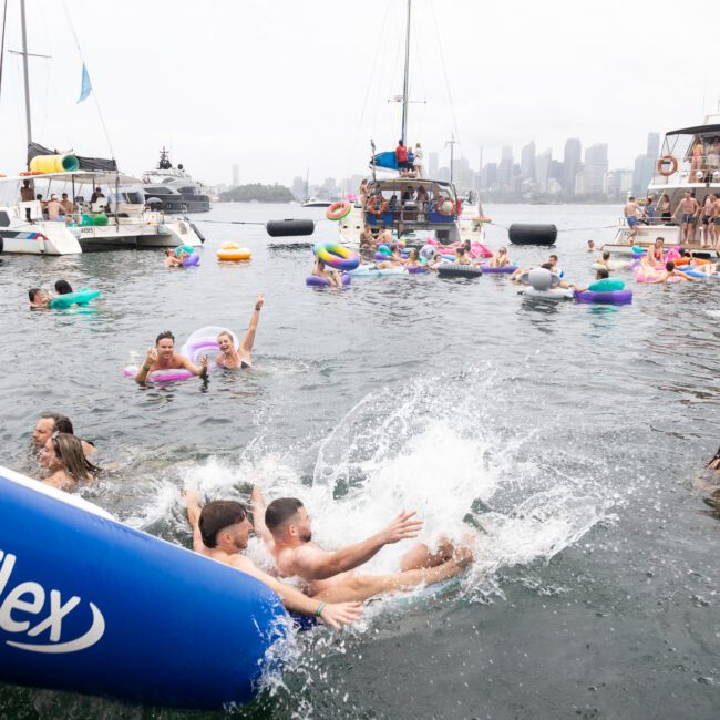 People are enjoying a boat party in a bay. Some are swimming, others are on inflatable pool floats, and a few are sliding from an inflatable slide. Several boats are anchored nearby, and a city skyline is visible in the misty background.