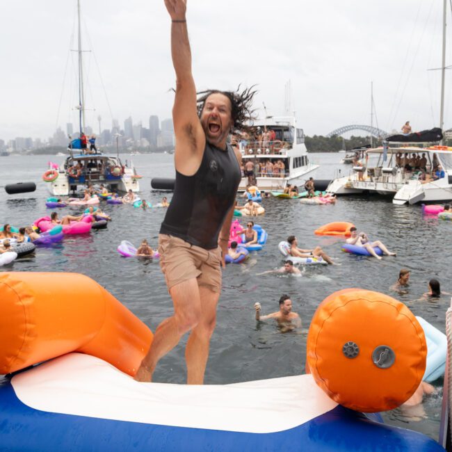 A man enthusiastically jumps off a large inflatable into the water, raising one arm in excitement. In the background, people relax on floaties and boats, with a city skyline visible under a cloudy sky.