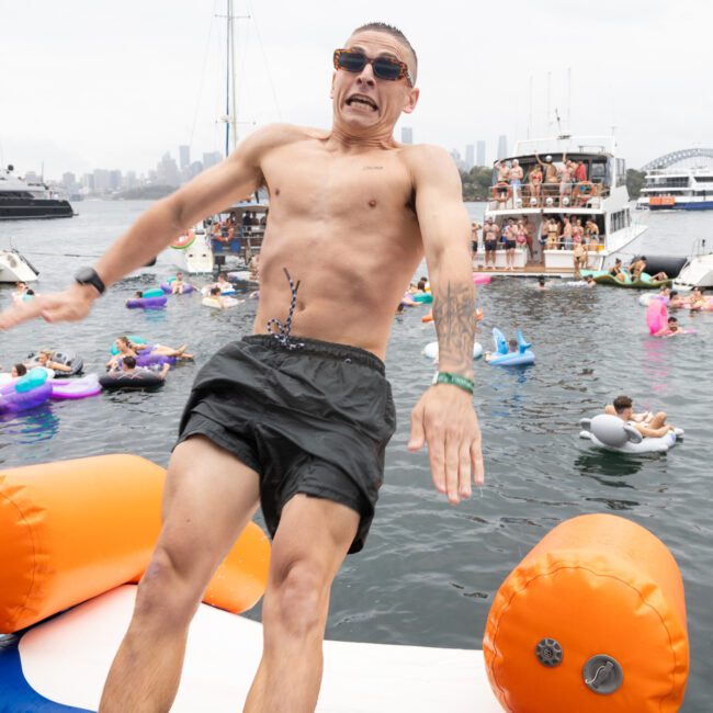 A man in black swim trunks and sunglasses is falling backward off an inflatable orange and white platform into the water. Boats and people in colorful pool floats are in the background, with a city skyline visible under a cloudy sky.