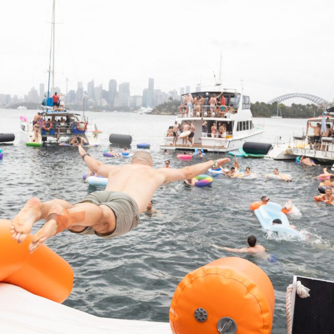 A person dives off an inflatable platform into the water, surrounded by people swimming and relaxing on floats. Several boats and yachts are in the background, with a city skyline and bridge visible in the distance. It appears to be a lively gathering.