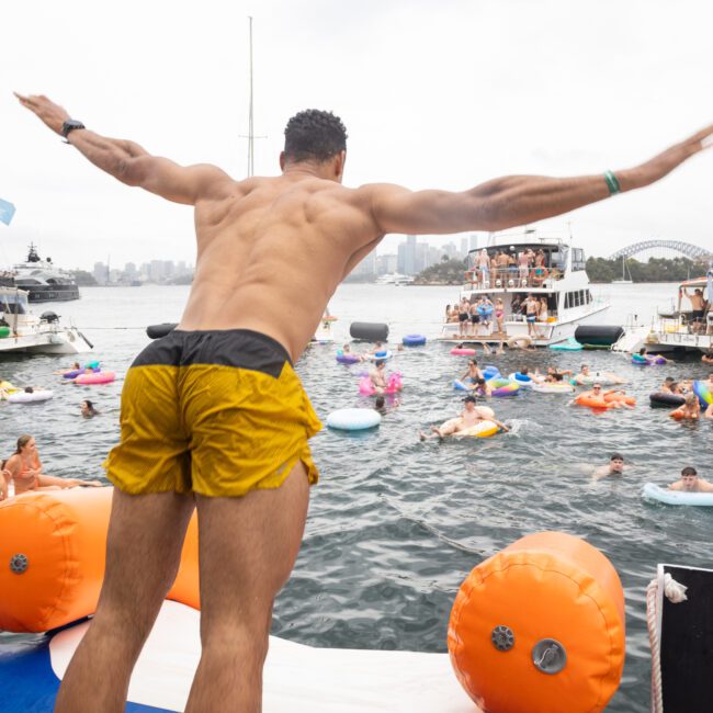 A man in yellow shorts prepares to dive into a body of water from a dock, surrounded by people enjoying a festive atmosphere on inflatable boats and floaties. Several boats are anchored nearby, and the city skyline is visible in the background.