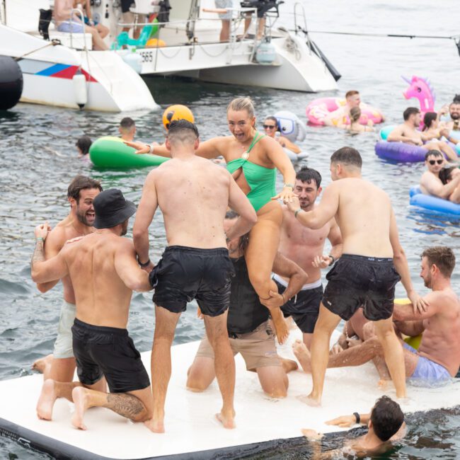 A group of people enjoy a lively day at the beach, gathered around a floating platform. Some are standing, others are on colorful inflatables in the water. Boats are visible in the background, creating a festive summer atmosphere.