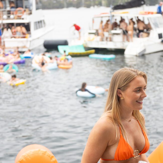 A woman in an orange bikini holds a can while smiling on a boat. Behind her, people enjoy the water on inflatables and other boats are nearby. A bridge and cityscape can be seen in the background.