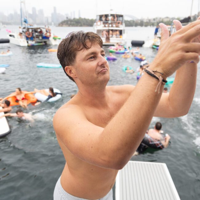 A shirtless man is standing on a boat deck, gesturing with his hands. Behind him, people are enjoying the water on floating devices and boats. The city skyline is visible in the misty background.