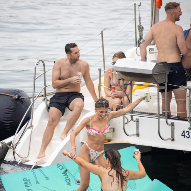 A group of people enjoy a day on a boat. Two women in swimsuits dance on a floating mat in the water beside the boat. A few others relax on the boat, with drinks in hand, under a cloudy sky.