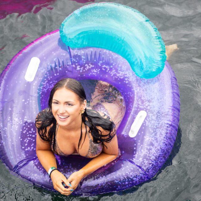 A woman in a swimsuit smiles while relaxing on a vibrant purple and blue inflatable float in the water. The overcast sky reflects on the water's surface. Water droplets are visible on the float.