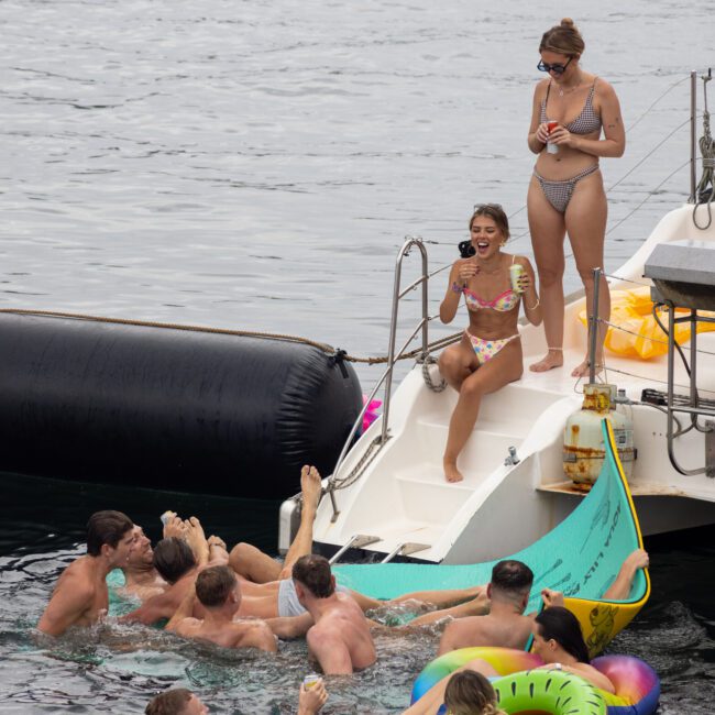 A group of people enjoying a boat party. Several individuals are swimming in the water near a slide attached to the boat. Others are standing or sitting on the boat deck, some holding drinks. The atmosphere is lively and festive.