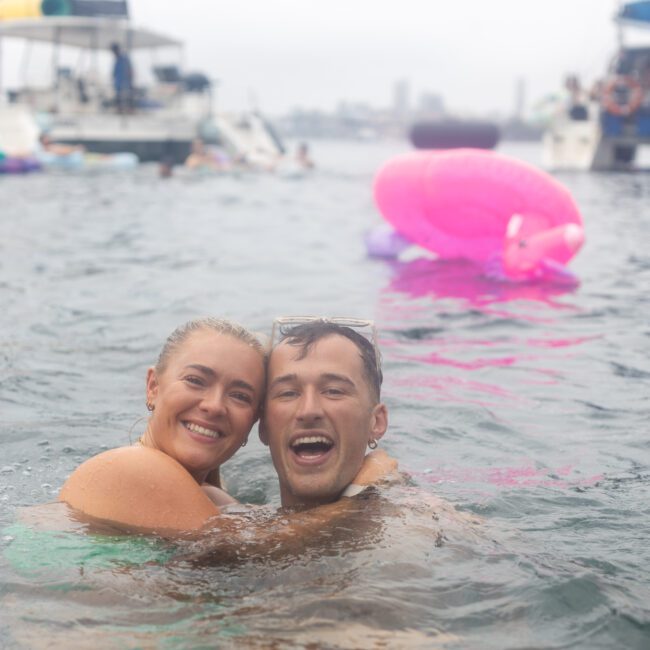 A couple smiling and embracing while swimming in the ocean. In the background, there are boats and a large pink inflatable flamingo float. The atmosphere is lively and joyful.