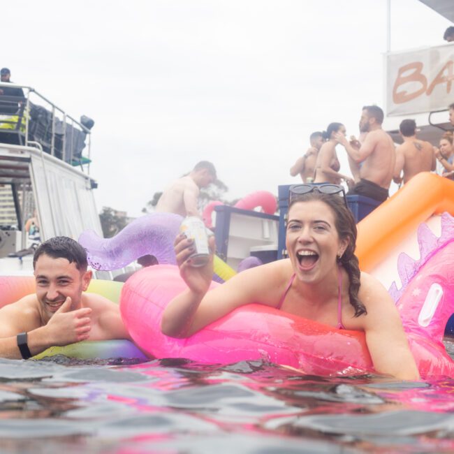 People enjoying a sunny day on the water. A woman smiles while holding a drink on a pink unicorn float, and a man gives a thumbs-up next to her. In the background, more guests socialize on a boat with a "Barefoot" sign.