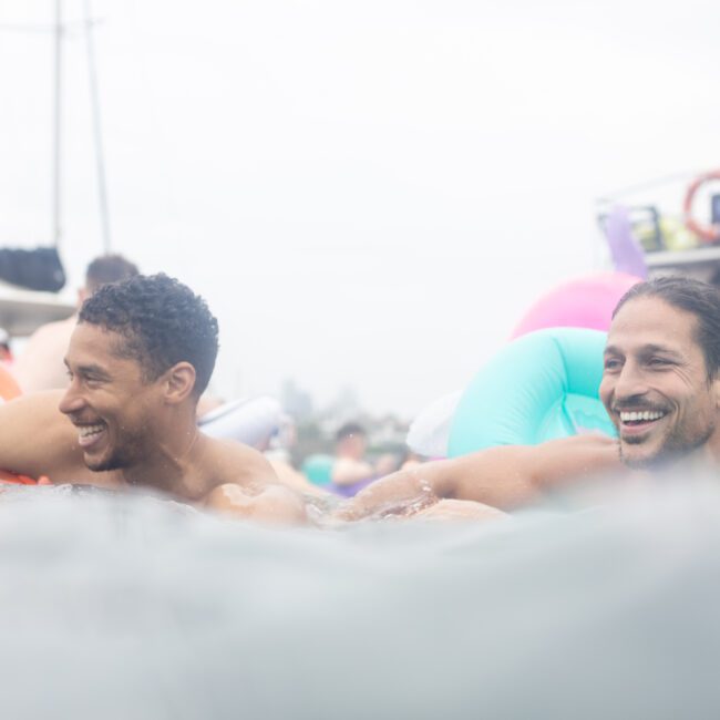 Two smiling men in the water, leaning on inflatable pool floats. The background shows a boat with people onboard. The atmosphere is joyful and relaxed, suggesting a fun day on the water.