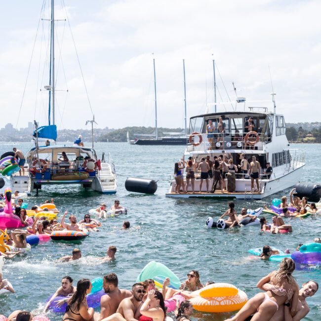 Groups of people enjoy a sunny day on the water, with several individuals relaxing on inflatables and a floating platform. Two boats are anchored in the background, hosting more people. The scene is festive and lively.