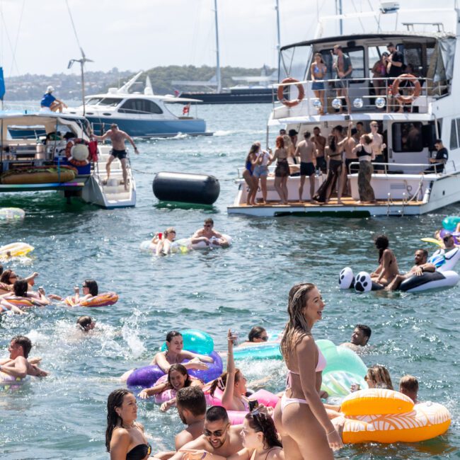 A lively scene with people swimming and relaxing on inflatables in a sunny bay. A group sits on a floating platform, while others are on boats in the background, enjoying the weather.