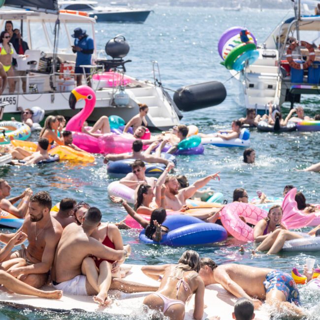 A lively scene of people enjoying a party on a lake, surrounded by boats. Many are on colorful inflatable floats, including a flamingo and unicorn. The atmosphere is festive, with people socializing and relaxing in the sun.