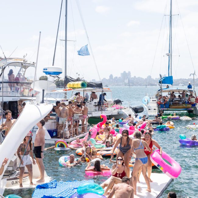 A lively scene on the water with numerous people enjoying a sunny day on inflatable rafts and boats. The crowd is having fun in bathing suits, surrounded by colorful floats. Sailboats can be seen in the background against a city skyline.