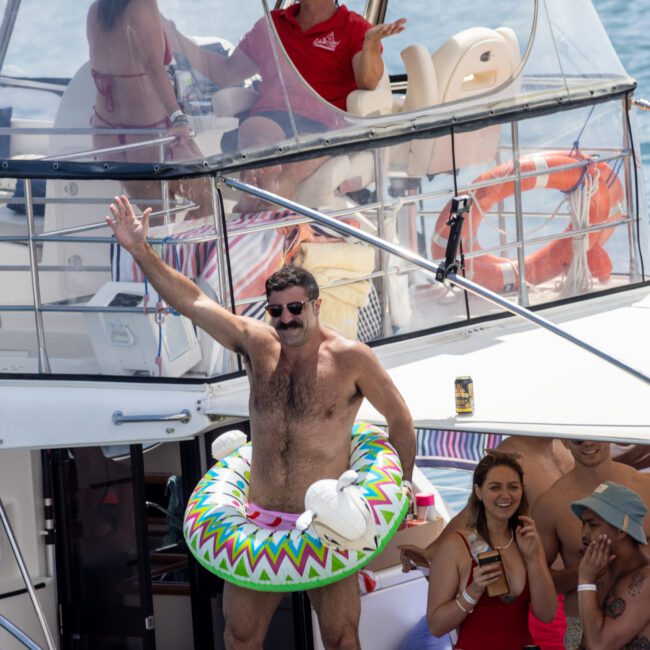 A man wearing a colorful inflatable ring gestures energetically, standing on a boat deck. Other people relax around him, some smiling and others talking. A man in a red shirt is seated on an upper deck. The scene is lively and joyous.