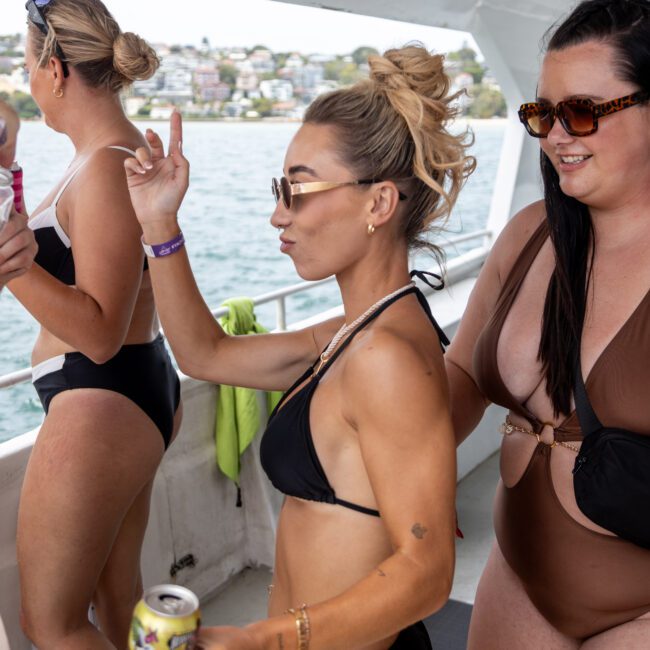 Three people in swimsuits are enjoying a boat ride. One person holds a can and seems to be dancing. The ocean and a coastline with houses are visible in the background.