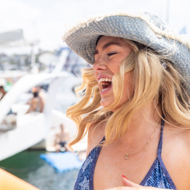 A woman wearing a straw hat and a blue patterned swimsuit laughs joyfully on a boat. The background shows other people on boats and water, creating a festive atmosphere under a sunny sky.