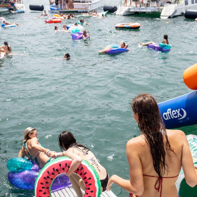 People enjoying a sunny day on the water with inflatable tubes and floaties. Boats are anchored nearby, and some individuals are diving in, while others relax on the floaties. In the foreground, a person holds a watermelon-shaped float.