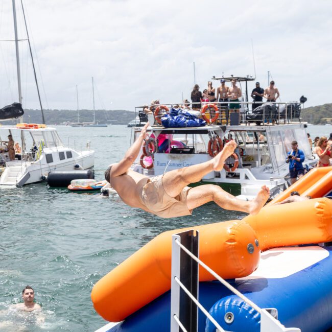 A person dives off a floating platform towards the water, surrounded by inflatable structures. People on nearby boats and in the water watch and cheer. The scene is lively, with multiple boats and people enjoying a sunny day on the water.