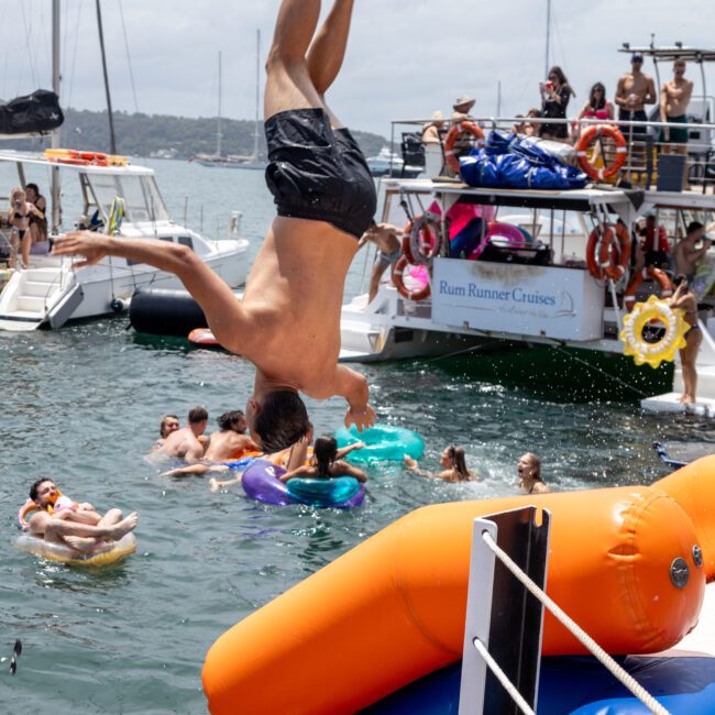 A person flipping into the water from an inflatable water slide on a boat. Several people are floating nearby, and other boats are in the background under a partly cloudy sky.