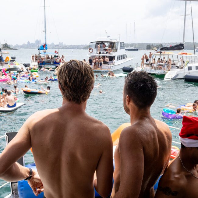 A crowded boat party with people swimming and relaxing on inflatable rafts. Two men stand at the edge of a boat looking at others enjoying the water. Boats are anchored nearby. One person is wearing a Santa hat. The atmosphere is lively and festive.