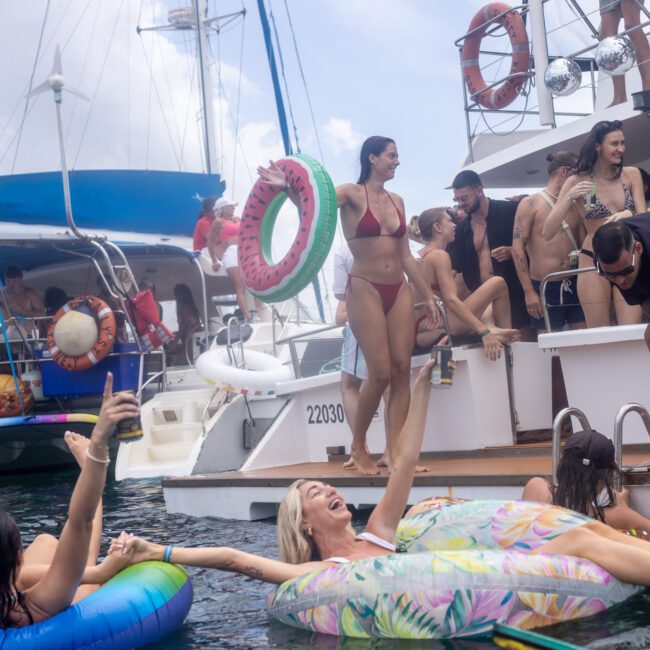 A group of people enjoying a yacht party. Several individuals are in swimsuits, socializing on the boat and in the water. One woman floats on an inflatable pool toy, and another holds a watermelon-themed inflatable ring. The sky is partly cloudy.