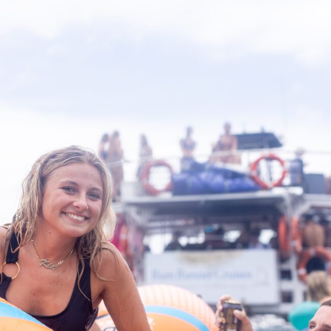 A woman smiles as she rests on an inflatable float in the water. In the background, a boat with people on board is visible, set against a bright sky.