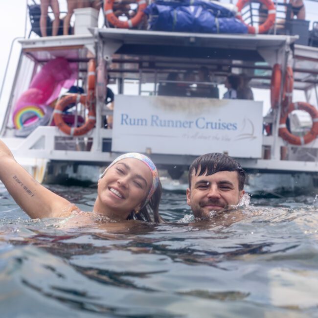 A man and woman smile while swimming in the water near a boat named "Rum Runner Cruises." The woman raises one arm in a playful gesture. People are visible on the boat's deck, and colorful inflatables can be seen behind them.