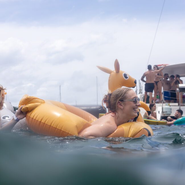 A woman in sunglasses smiles while floating on an inflatable kangaroo in the water. Other people are in the background near boats, enjoying the sunny day.